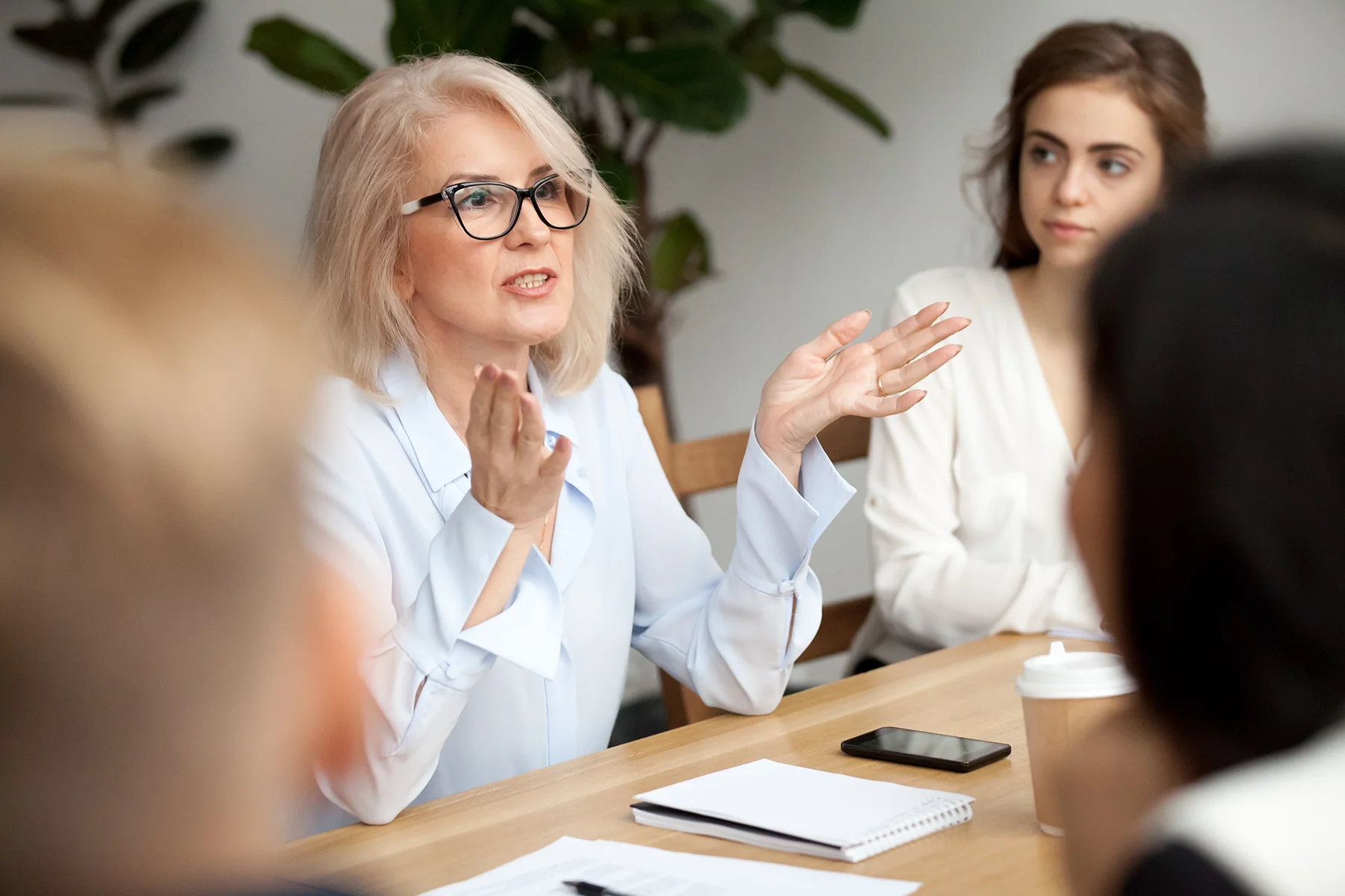 Weibliche Führungskraft sitzt mit ihrem Team am Tisch und spricht in die Runde – Erika Götz, Führungskräftecoaching